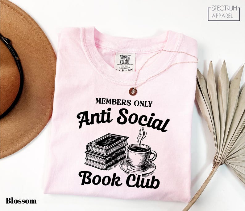 a pink t-shirt with a graphic design that says "MEMBERS ONLY Anti Social Book Club" and includes a book and a cup of coffee. The t-shirt is laid out on a white surface, with a brown hat and a plant visible in the background.