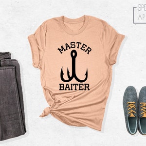 Pode incluir: Uma camiseta cor de p&ecirc;ssego com um gr&aacute;fico preto de um anzol de pesca e o texto "Master Baiter".