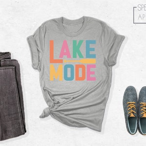 May include: A heather grey t-shirt with the text "LAKE MODE" in colorful letters. The letters are in different colors, including orange, pink, yellow, blue, and green. A small orange paddle is between the words "LAKE" and "MODE".