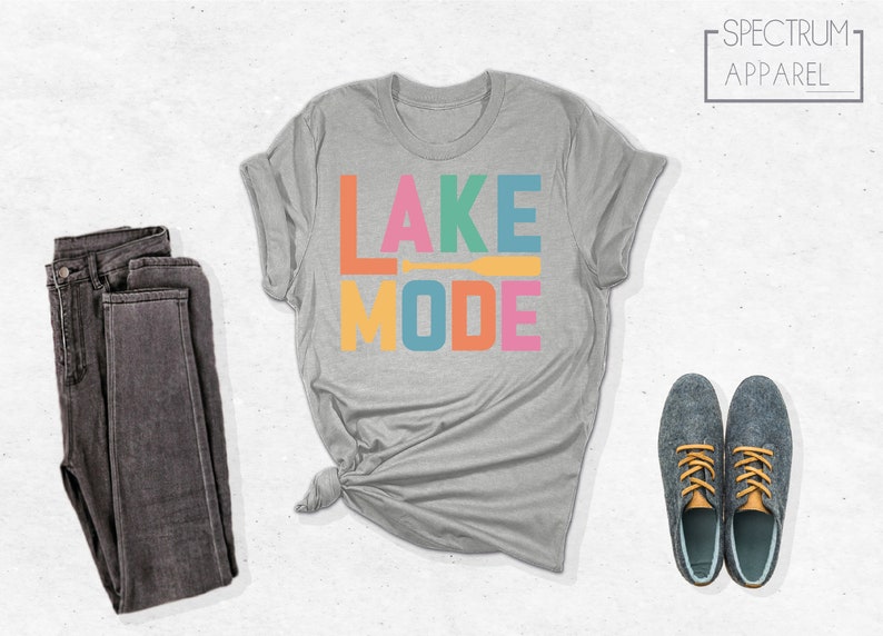 May include: A heather grey t-shirt with the text "LAKE MODE" in colorful letters. The letters are in different colors, including orange, pink, yellow, blue, and green. A small orange paddle is between the words "LAKE" and "MODE".