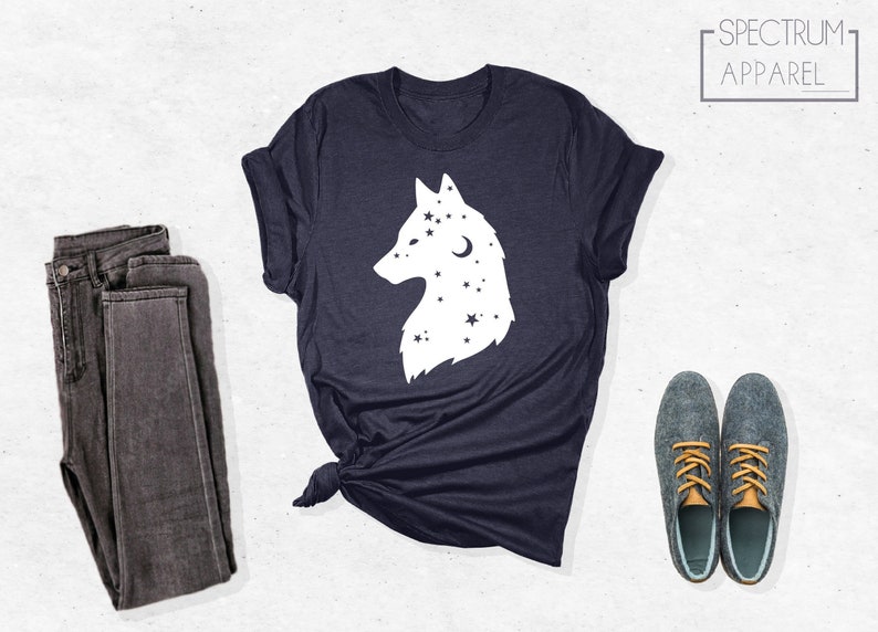 May include: A navy blue t-shirt with a white wolf silhouette design. The wolf is surrounded by stars and a crescent moon. The t-shirt is folded and laid flat on a white surface. The text "SPECTRUM APPAREL" is in a white rectangle in the upper right corner of the image.