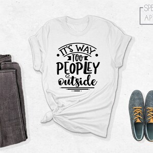 May include: White t-shirt with black text that reads "It's Way Too Peopley Outside". The shirt is folded and laid flat on a white surface. There are a pair of dark gray jeans and two gray shoes with orange laces on the surface.
