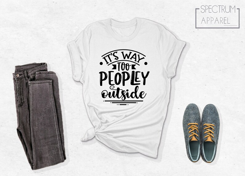 May include: White t-shirt with black text that reads "It's Way Too Peopley Outside". The shirt is folded and laid flat on a white surface. There are a pair of dark gray jeans and two gray shoes with orange laces on the surface.