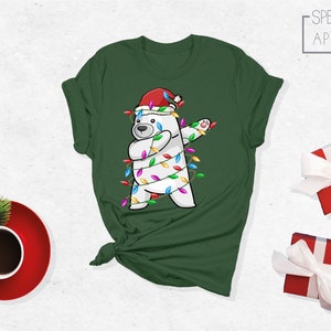 Op de afbeelding: Groen t-shirt met een witte ijsbeer met een kerstmuts en omwikkeld met kerstverlichting. De beer doet een dab. Het shirt heeft het Spectrum Apparel logo.