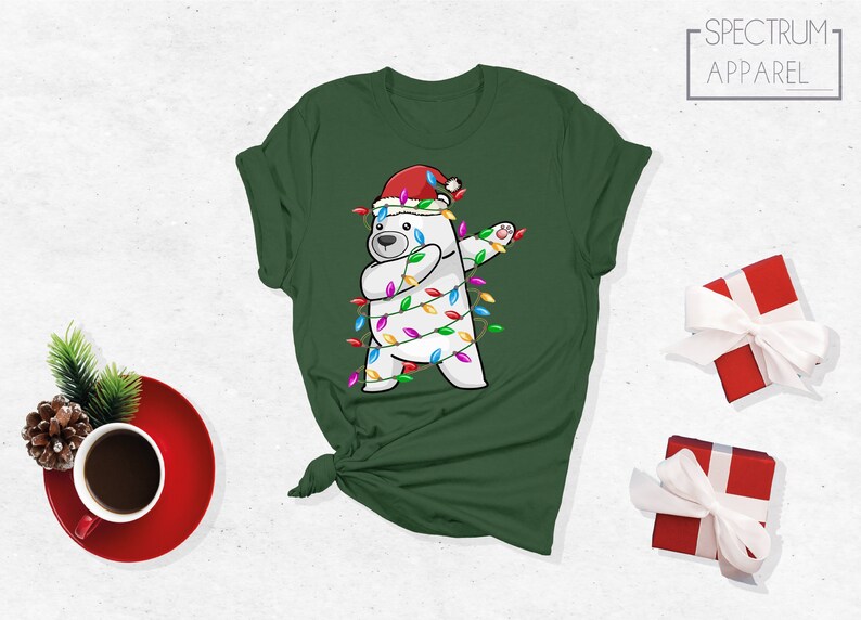 Op de afbeelding: Groen t-shirt met een witte ijsbeer met een kerstmuts en omwikkeld met kerstverlichting. De beer doet een dab. Het shirt heeft het Spectrum Apparel logo.