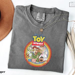 a gray t-shirt with a colorful graphic of characters from the Toy Story movie, including Woody, Buzz Lightyear, and other beloved characters. The t-shirt is laid out on a white surface, with a brown hat and some leaves visible in the background.