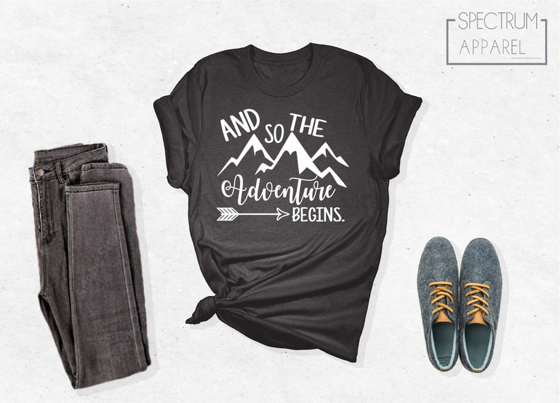 May include: A dark gray t-shirt with a white graphic design that says "And so the adventure begins." with a mountain range and an arrow pointing to the right.
