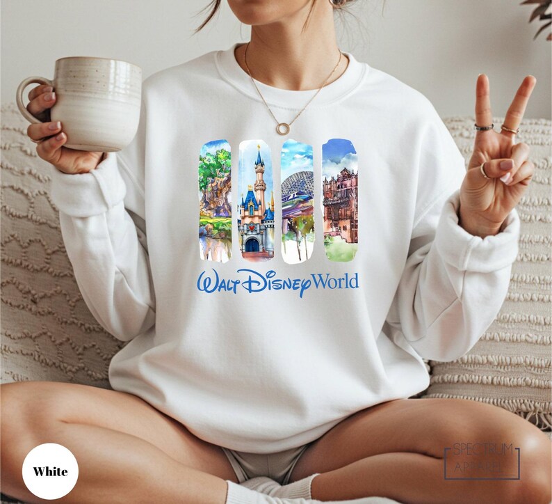 A woman wearing a white sweater with a collage of Disney World images and the text " Walt Disney World" printed on it, holding a mug and making a peace sign.