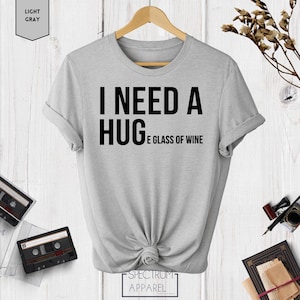 May include: A light gray t-shirt with the text "I need a hug and a glass of wine" printed in black.