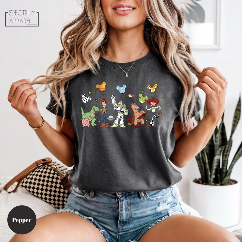 A woman wearing a black t-shirt with a graphic of various cartoon characters, including a dog, a pig, and a cow, along with a necklace. She is sitting in a room with a potted plant and a checkered bag.