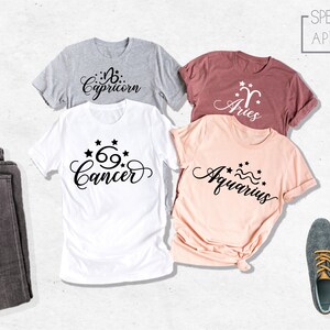 May include: Four t-shirts with different zodiac signs printed on them. The signs are Capricorn, Aries, Cancer, and Aquarius. The shirts are in different colors, including gray, pink, and maroon. The shirts are laid out on a white surface.