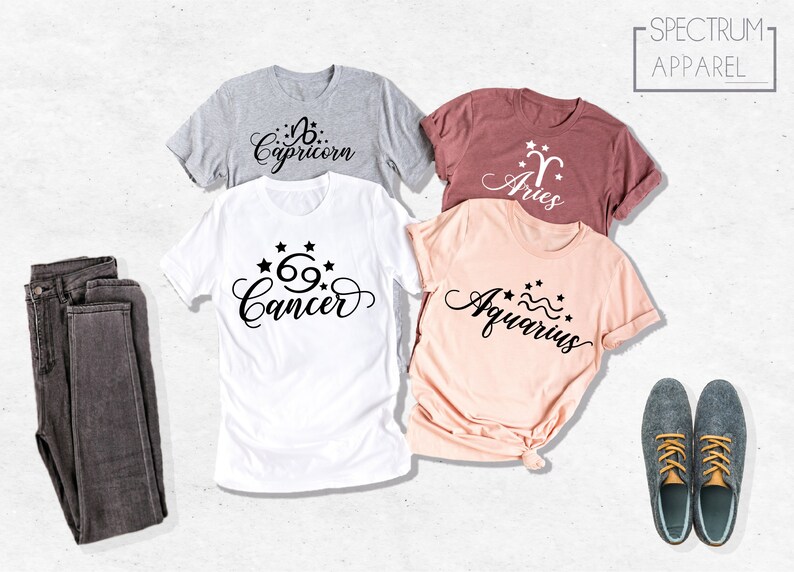 May include: Four t-shirts with different zodiac signs printed on them. The signs are Capricorn, Aries, Cancer, and Aquarius. The shirts are in different colors, including gray, pink, and maroon. The shirts are laid out on a white surface.