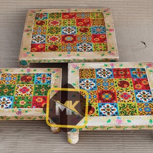 May include: Three square wooden stools with intricate painted designs. The tops of the stools are decorated with colorful geometric patterns and floral motifs. The stools are made of light wood and have a distressed finish.