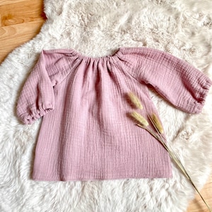 Könnte beinhalten: Ein staubiges rosafarbenes, langärmliges,  Gaze-Babykleid mit gerafftem Ausschnitt. Das Kleid liegt flach auf einer weißen Kunstfell-Oberfläche.