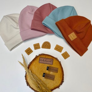 Könnte beinhalten: Fünf Waffelstrickmützen in verschiedenen Farben, darunter Creme, Rosa, Hellblau und Rost. Die Mützen sind auf einer weißen Oberfläche mit mehreren kleinen braunen Leder-Etiketten mit Text angeordnet, darunter "Ciao Kakao", "Hello World", "Hip Hip Hurra", "Frech Dachs", "Brother", "Sister" und "Wirbelwind".
