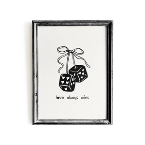 May include: Black and white framed art print featuring two dice hanging from a bow. The dice are decorated with hearts and other shapes. The text "love always wins" is written below the dice.