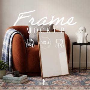 May include: A vertical wooden picture frame mockup with a blank white center, positioned in front of a rust-colored armchair with a blue plaid throw blanket, a grey pillow, a small ghost-shaped lamp, and books labeled Nordic Living and Eclectic Homes.