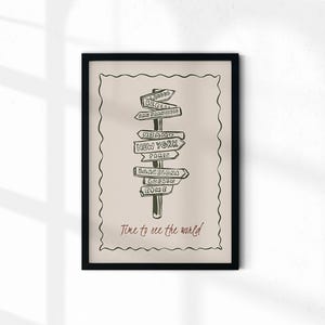 Puede incluir: Impresión artística enmarcada en negro con una ilustración dibujada a mano de un poste indicador con flechas que señalan ciudades como Londres, Nueva York y Roma. El texto "Time to see the world" está escrito en cursiva en la parte inferior.