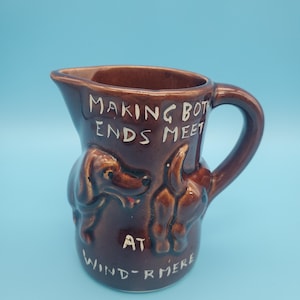 May include: A brown ceramic jug with a handle, featuring embossed dog illustrations and the text "MAKING BOTH ENDS MEET AT WIND-RMERE" in white lettering. The jug has a spout for pouring.