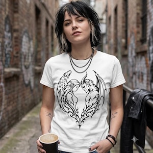May include: A white short-sleeved t-shirt featuring a black graphic print on the chest. The design depicts an angel with feathered wings facing a demon with horns and bat-like wings, their silhouettes forming a heart shape in the center.
