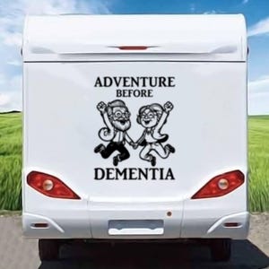 May include: White camper van with a black and white sticker on the back. The sticker shows a cartoon couple jumping and holding hands. The text on the sticker reads "Adventure Before Dementia".