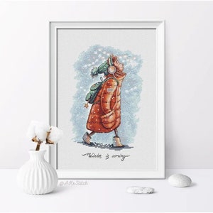 Peut inclure: Un motif de point de croix repr&eacute;sentant une fille portant une veste rouge et un chapeau vert marchant dans la neige. Le texte "Winter is coming" est sous la fille. Le motif est encadr&eacute; et sur une surface blanche.