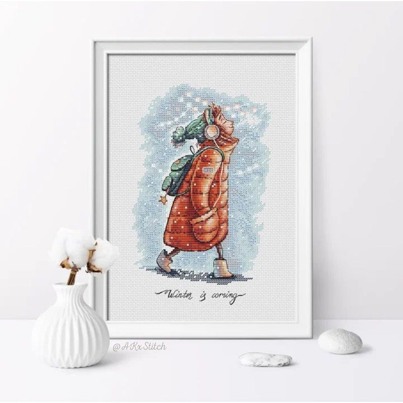Peut inclure: Un motif de point de croix repr&eacute;sentant une fille portant une veste rouge et un chapeau vert marchant dans la neige. Le texte "Winter is coming" est sous la fille. Le motif est encadr&eacute; et sur une surface blanche.