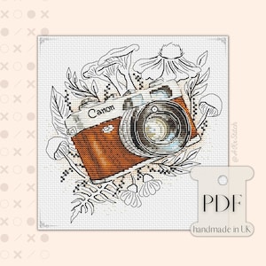 Puede incluir: Un patrón de punto de cruz que presenta una cámara vintage con la palabra "Canon" en ella, rodeada de flores y hongos estilizados. El patrón está en blanco y negro con una cámara marrón.