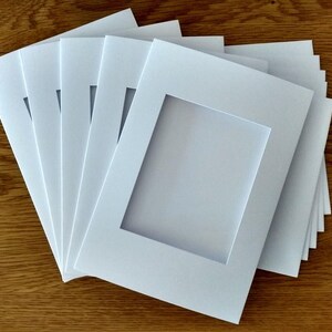 May include: A stack of white photo mats with a rectangular opening in the center. The mats are made of a thick, sturdy cardstock and are perfect for framing photos or artwork.