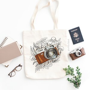 May include: A white canvas tote bag with a brown and black vintage camera illustration surrounded by flowers and mushrooms. The camera has the word "Canon" printed on it. The bag has a black handle.
