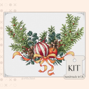 Könnte beinhalten: Ein Weihnachts-Kreuzstich-Set mit einem Kranz aus Kiefernästen, roten Beeren, Tannenzapfen und einem rot-weiß gestreiften Ornament mit einer gelben Schleife. Das Set ist handgefertigt in Großbritannien.