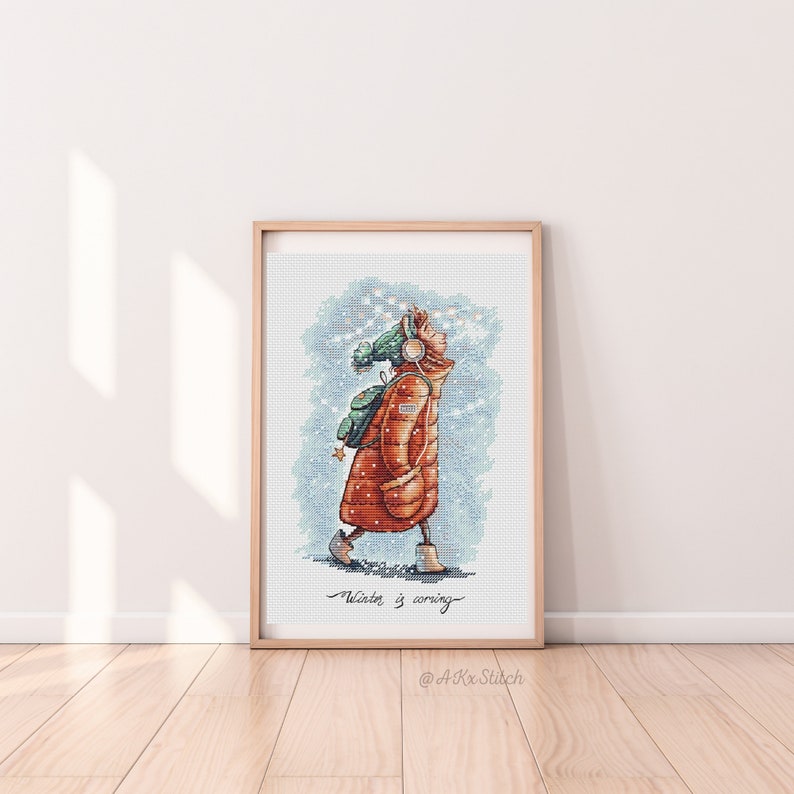 Peut inclure: Un motif de point de croix repr&eacute;sentant une personne portant un manteau d'hiver rouge et marron, un chapeau vert et des &eacute;couteurs, marchant dans la neige. Le texte "Winter is coming" est &eacute;crit sous la personne. Le motif est encadr&eacute; dans un cadre en bois clair.