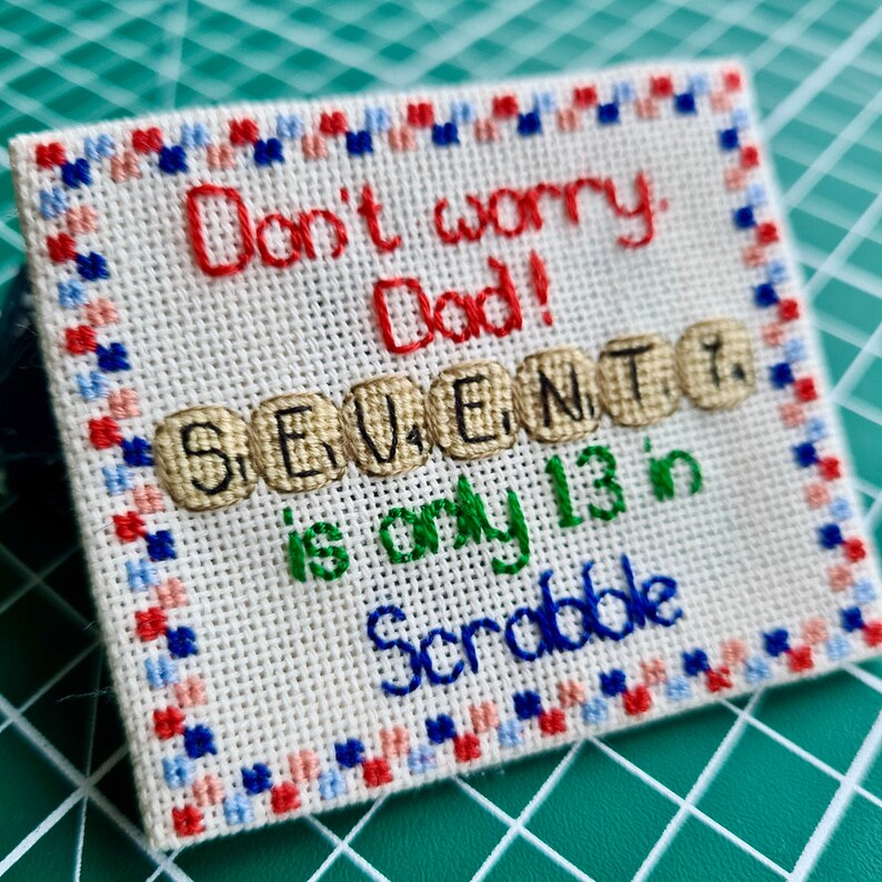 K&ouml;nnte beinhalten: Ein Kreuzstich-St&uuml;ck Stoff mit rotem Rand und wei&szlig;em Hintergrund. Der Text "Don't worry Dad! SEVEN is only 13 in Scrabble" ist in roter, gr&uuml;ner und schwarzer Farbe gestickt.