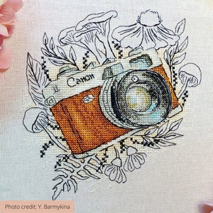May include: A cross-stitch embroidery of a vintage camera with the word "Canon" on the front. The camera is surrounded by black and white outlines of flowers, mushrooms, and leaves.