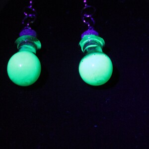 May include: A pair of glowing green earrings against a black background. Each earring features a spherical, luminous green bead, a clear, wavy accent, and a purple bead. The earrings are suspended from a silver-colored, swirled hook.