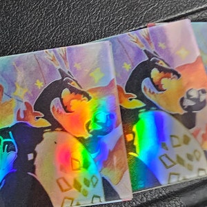 May include: Three iridescent holographic stickers with a colorful abstract design featuring a black silhouette of a creature with a crown. The background is a light purple with white stars.