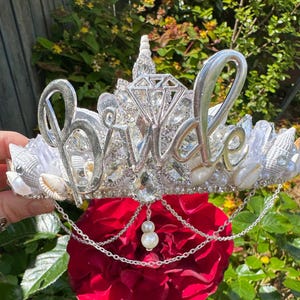 May include: A silver tiara with the word "Bride" in cursive script, adorned with crystals, seashells, and pearls. A large diamond-shaped crystal sits at the center. A red rose is below the tiara, with a chain and pearl pendant.
