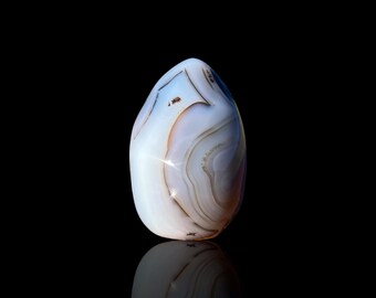 May include: A smooth, oval gemstone with a pattern of white, tan, and brown bands. The stone is polished and reflects light, set against a black background. The stone's reflection is visible below.