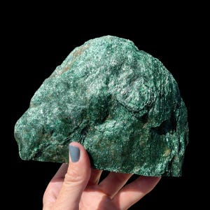 May include: A large, raw chunk of green fuchsite mineral with a highly reflective, crystalline surface texture. The stone displays deep emerald and forest green hues with natural fissures and a jagged, uneven shape throughout the specimen.