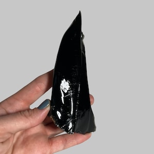 May include: A polished, black obsidian stone with a glossy, reflective surface. The irregularly shaped stone is held in a hand against a neutral background. The obsidian has a sharp, pointed top and a rough, textured base.