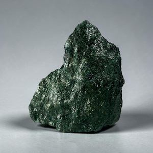 May include: A raw, unpolished mineral specimen with a deep forest green color. The stone features a jagged, uneven surface with a crystalline, reflective texture that catches the light, creating a multifaceted appearance across its irregular shape.