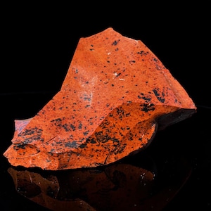 May include: A raw, natural obsidian stone with a vibrant orange hue and speckled with black inclusions. The rough, irregular shape of the stone is displayed against a black background, highlighting its unique texture and color variations. A striking example of natural mineral beauty.