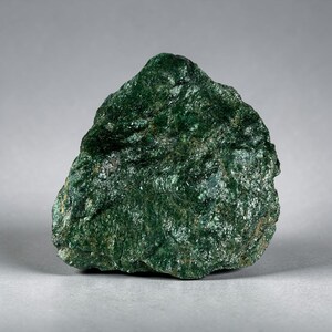May include: A raw, unpolished mineral specimen featuring a deep forest green color with a crystalline, textured surface. The stone has an irregular, jagged shape with visible light-reflecting facets across its dense, dark green, rocky composition.