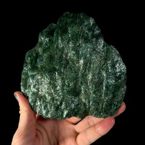 May include: A raw, unpolished mineral specimen featuring a deep forest green color with a highly reflective, crystalline surface texture. The stone has an irregular, jagged shape with visible mica-like flakes that catch the light across its surface.