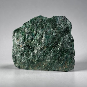 May include: A raw, unpolished mineral specimen featuring a deep forest green color with a highly textured, crystalline surface. The stone displays natural variations in shade, ranging from dark emerald to blackish green, with a reflective, shimmering quality across its jagged, uneven edges and dense, compact structure.