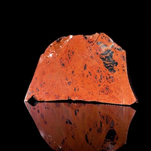 May include: A rough-cut, reddish-orange obsidian stone with black inclusions. The stone is displayed against a black background, with its reflection visible below. The natural stone has a textured surface and irregular edges.