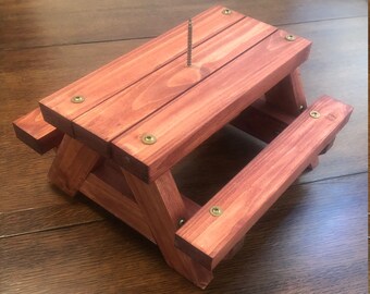 May include: A miniature wooden picnic table with a red-brown stain finish. The table has a small screw in the center of the tabletop.
