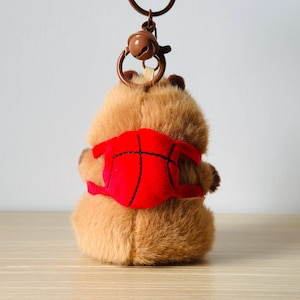 Op de afbeelding: Een bruine pluche dieren-sleutelhanger met een kleine, ronde, rode rugzak met zwarte stiksels die op een basketbal lijken. De sleutelhanger is voorzien van een bruine metalen ring en een klein bruin belletje aan de bovenkant van het figuurtje.