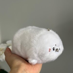 May include: A white plush seal toy with a small silver keychain attached. The seal has a round body, small black eyes, and pink blush marks on its cheeks. The toy is made of soft, fluffy material.