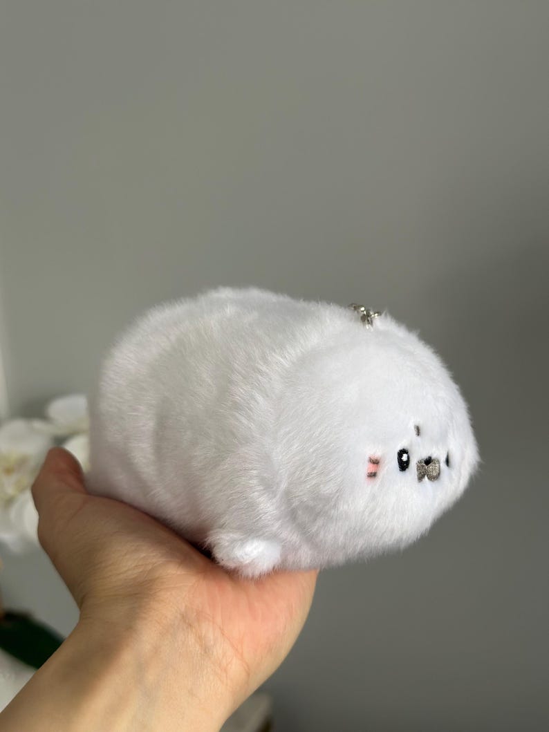 May include: A white plush seal toy with a small silver keychain attached. The seal has a round body, small black eyes, and pink blush marks on its cheeks. The toy is made of soft, fluffy material.